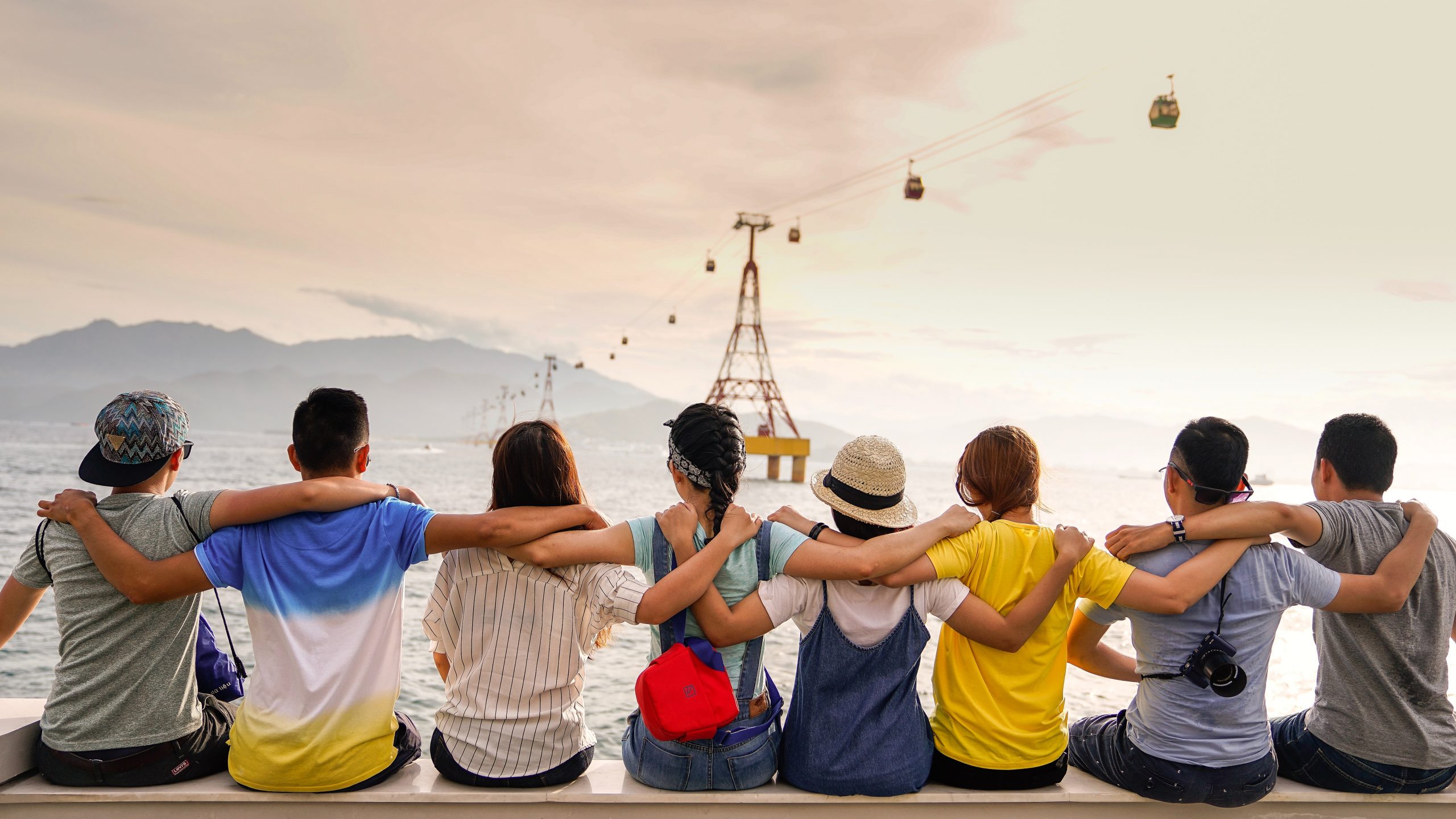 Um grupo de amigos da República Estudantil sentados à beira-mar, observando os teleféricos suspensos sobre o mar, tendo como pano de fundo montanhas distantes.