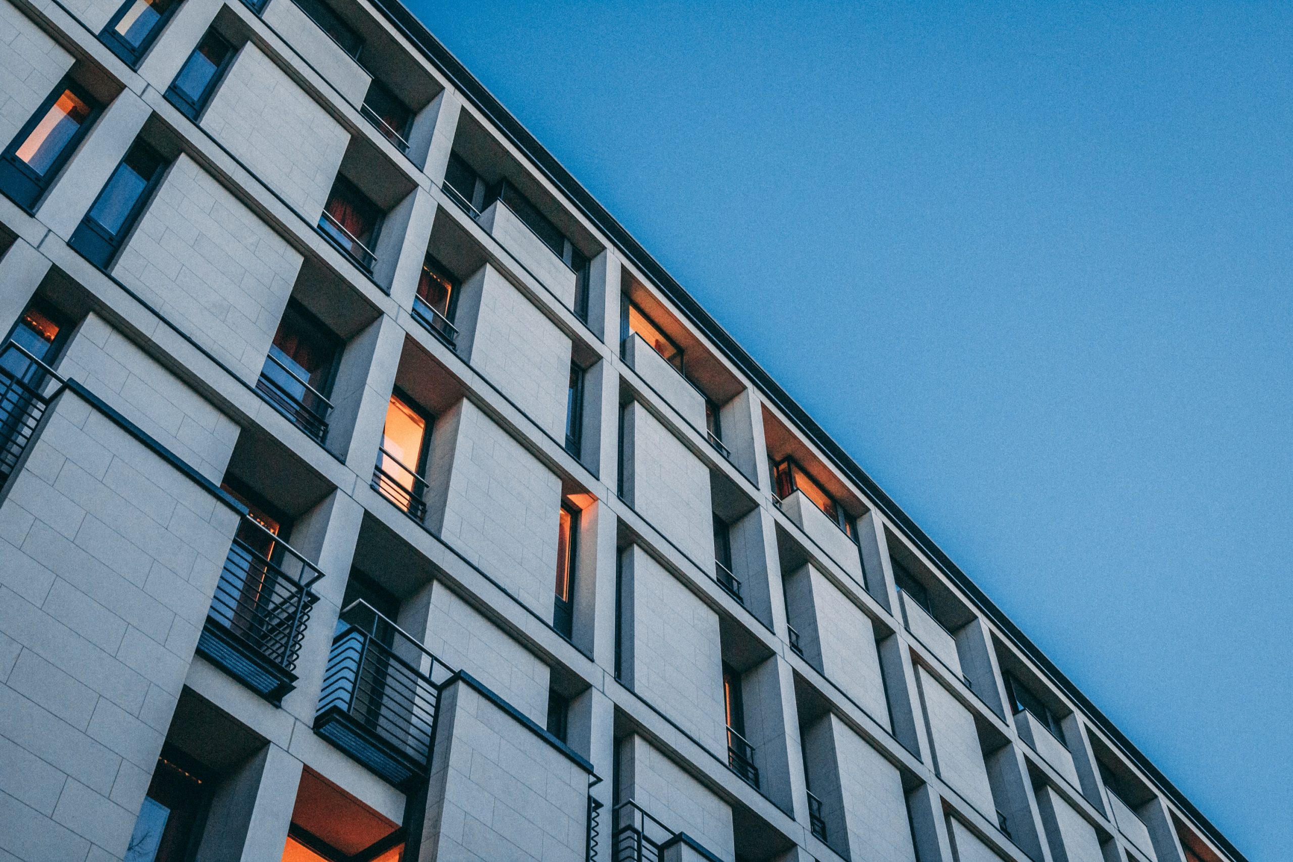Edifício moderno de alojamento estudantil ao entardecer, com o brilho quente das luzes visíveis de algumas das janelas contra o azul frio do céu noturno.