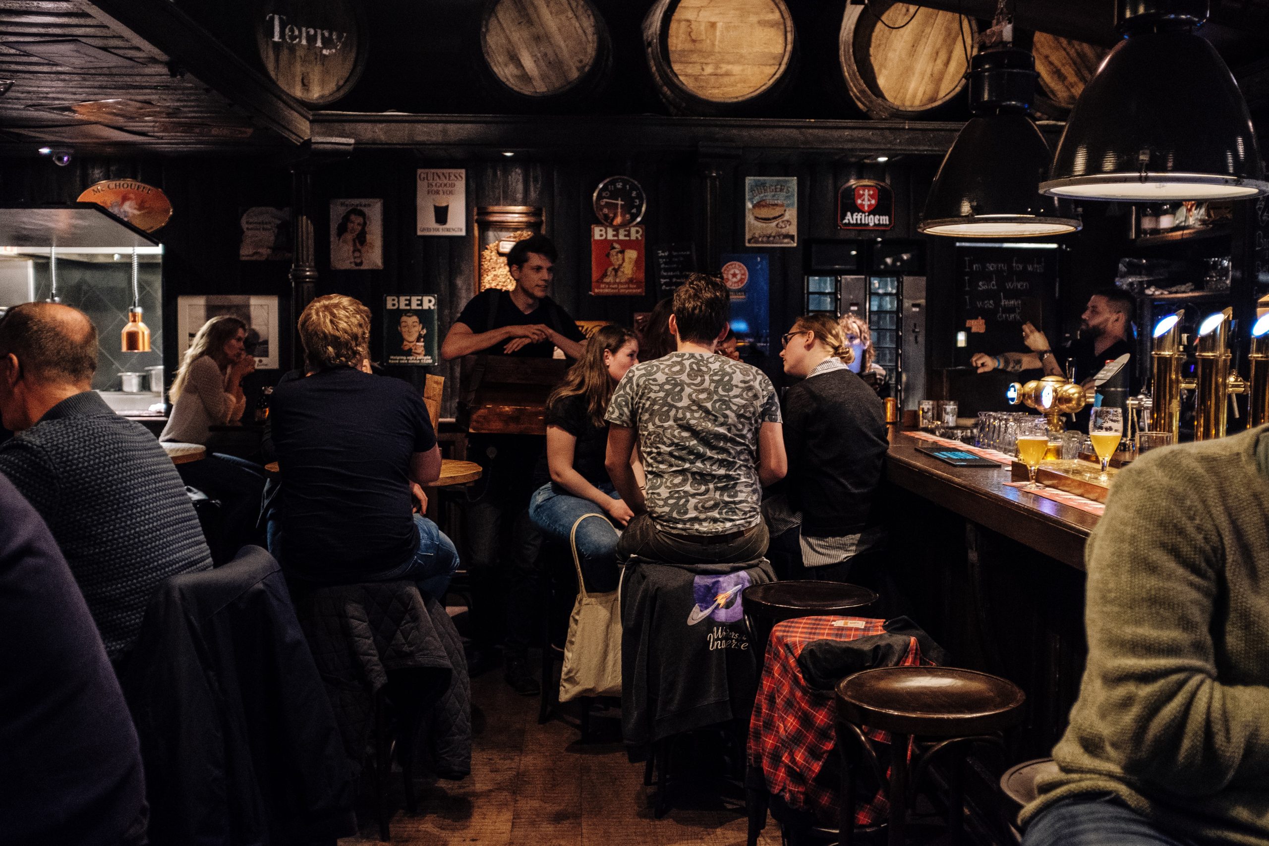 Clientes desfrutando de uma noite aconchegante em um pub universitário mal iluminado com um barman servindo bebidas.