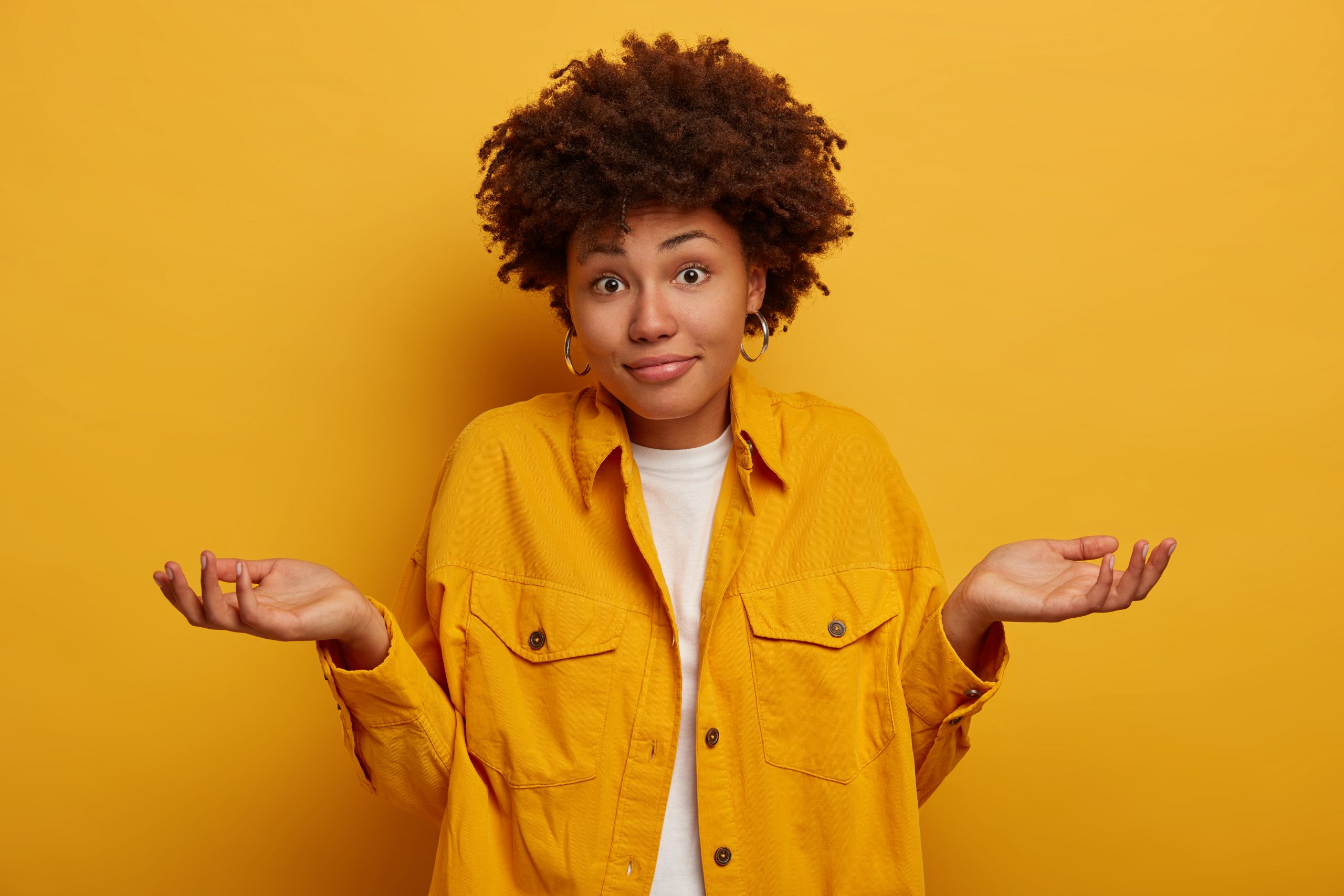 puzzled-clueless-african-american-lady-shrugs-shoulders-and-expresses-uncertainty-makes-decision-wears-fashionable-clothing-spreads-palms-sideways-isolated-on-yellow-wall-life-perception-attitude