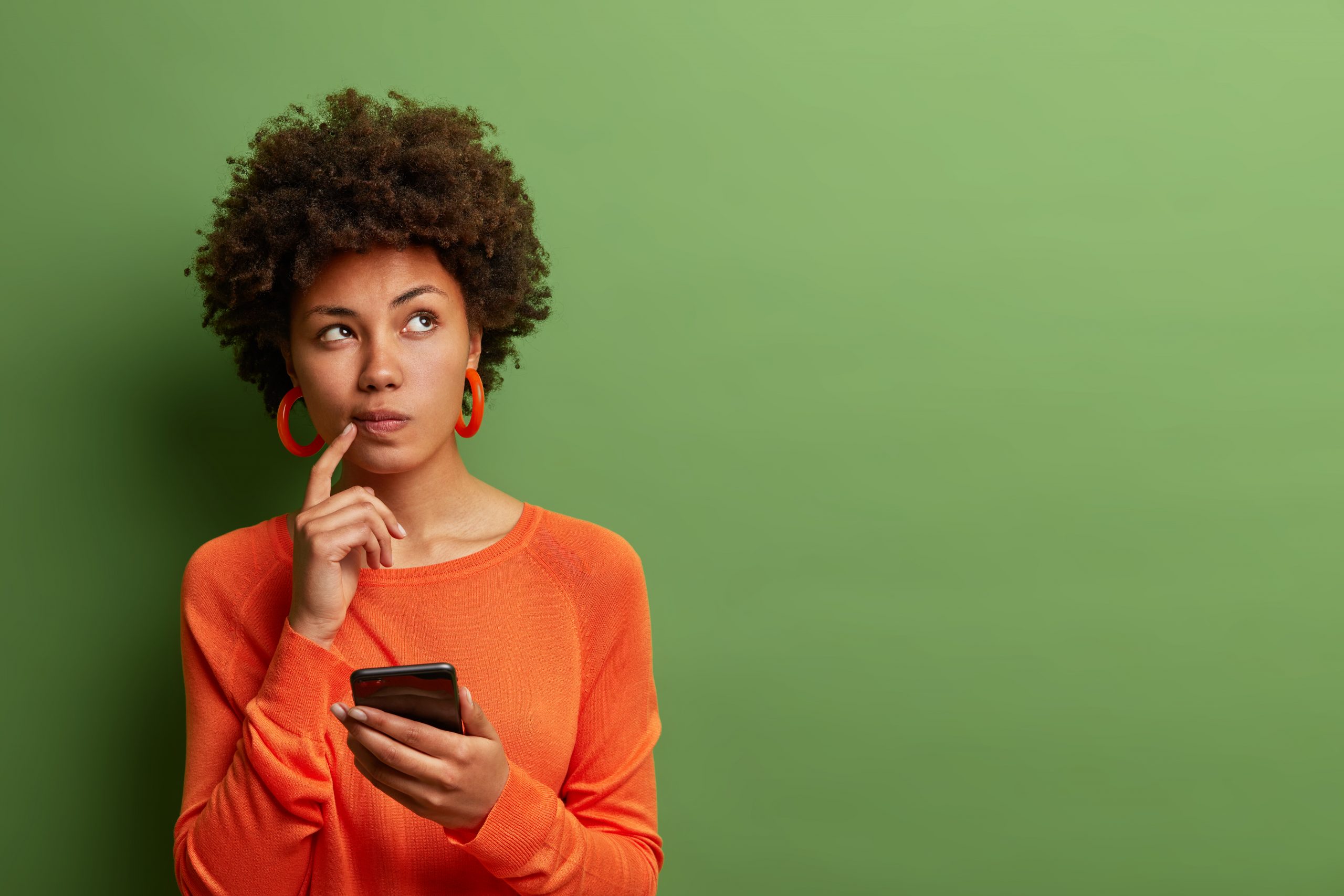 photo-of-pretty-ethnic-woman-ponders-on-how-to-answer-question-thinks-deeply-about-something-uses-modern-mobile-phone-tries-to-made-up-good-message-keeps-index-finger-near-lips-stands-indoor
