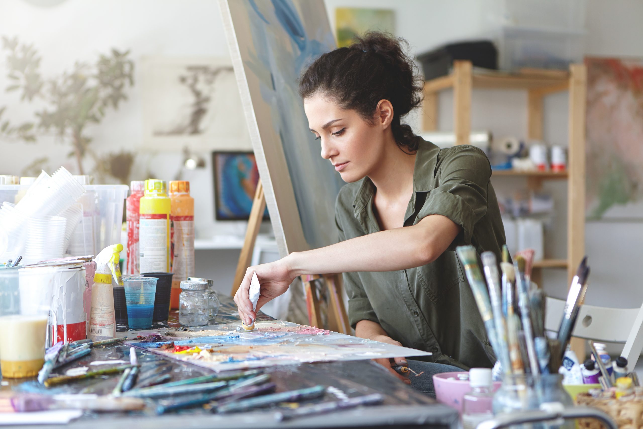 serious-brunette-young-beautiful-woman-sitting-in-art-studio-taking-colorful-paints-from-tube-while-creating-great-masterpiece-on-easel-being-preoccupied-with-her-work-having-nice-imagination