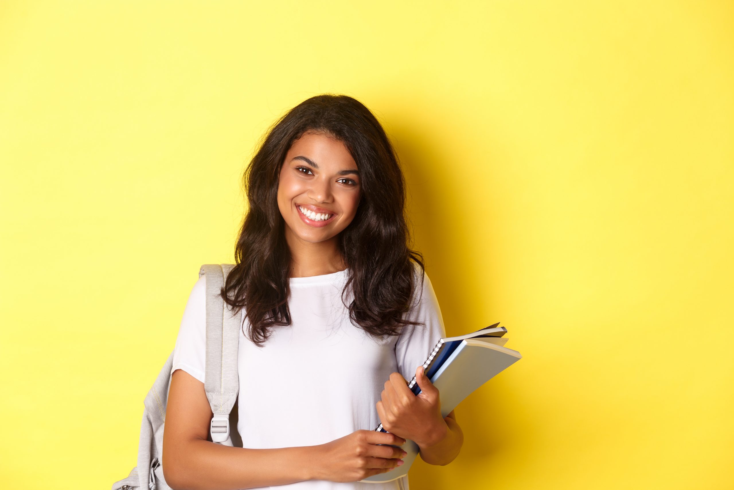 portrait-of-happy-african-american-female-college-student-holding-notebooks-and-backpack-smiling-and-standing-over-yellow-background