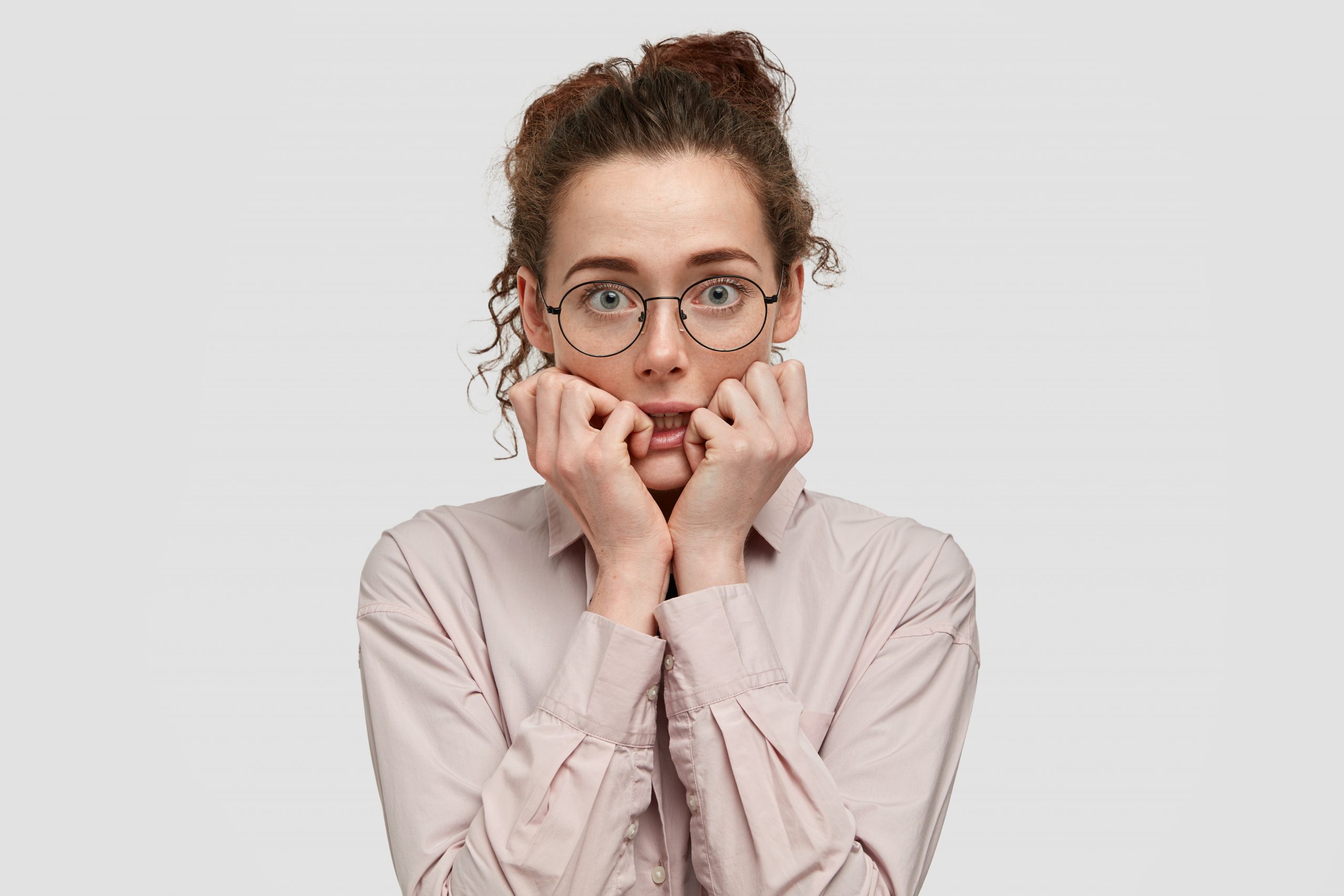 unhappy-nervous-young-female-with-worried-expression-bites-finger-nails-looks-anxiously-directly-at-camera-wears-spectacles-dressed-in-fashionable-clothes-stands-against-white-background