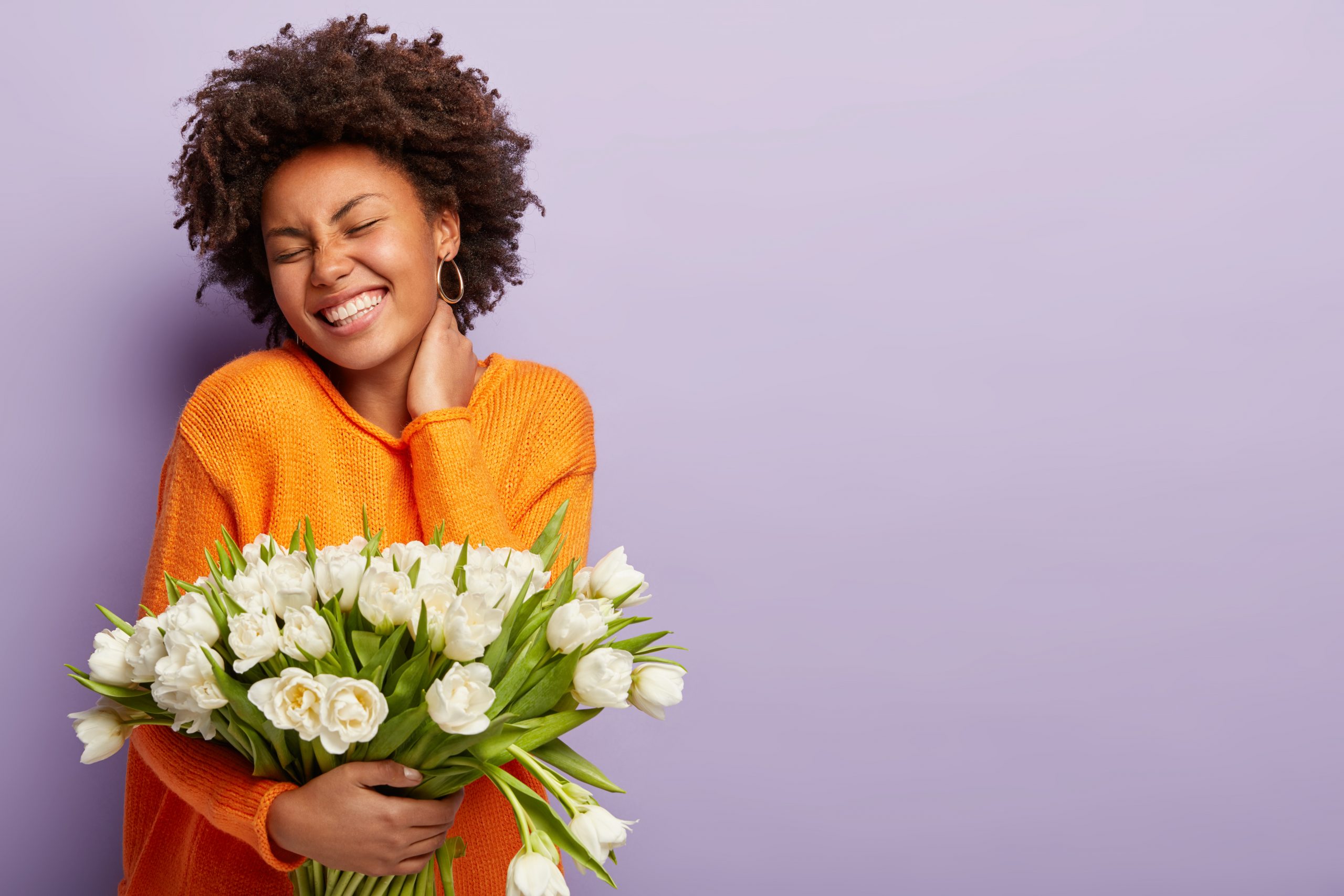 jubilant-upbeat-black-woman-has-curly-hair-broad-shining-smile-keeps-hand-on-neck-eyes-shut-wears-orange-sweater-holds-white-flowers-with-pleasant-scent-models-over-purple-studio-wall-spring